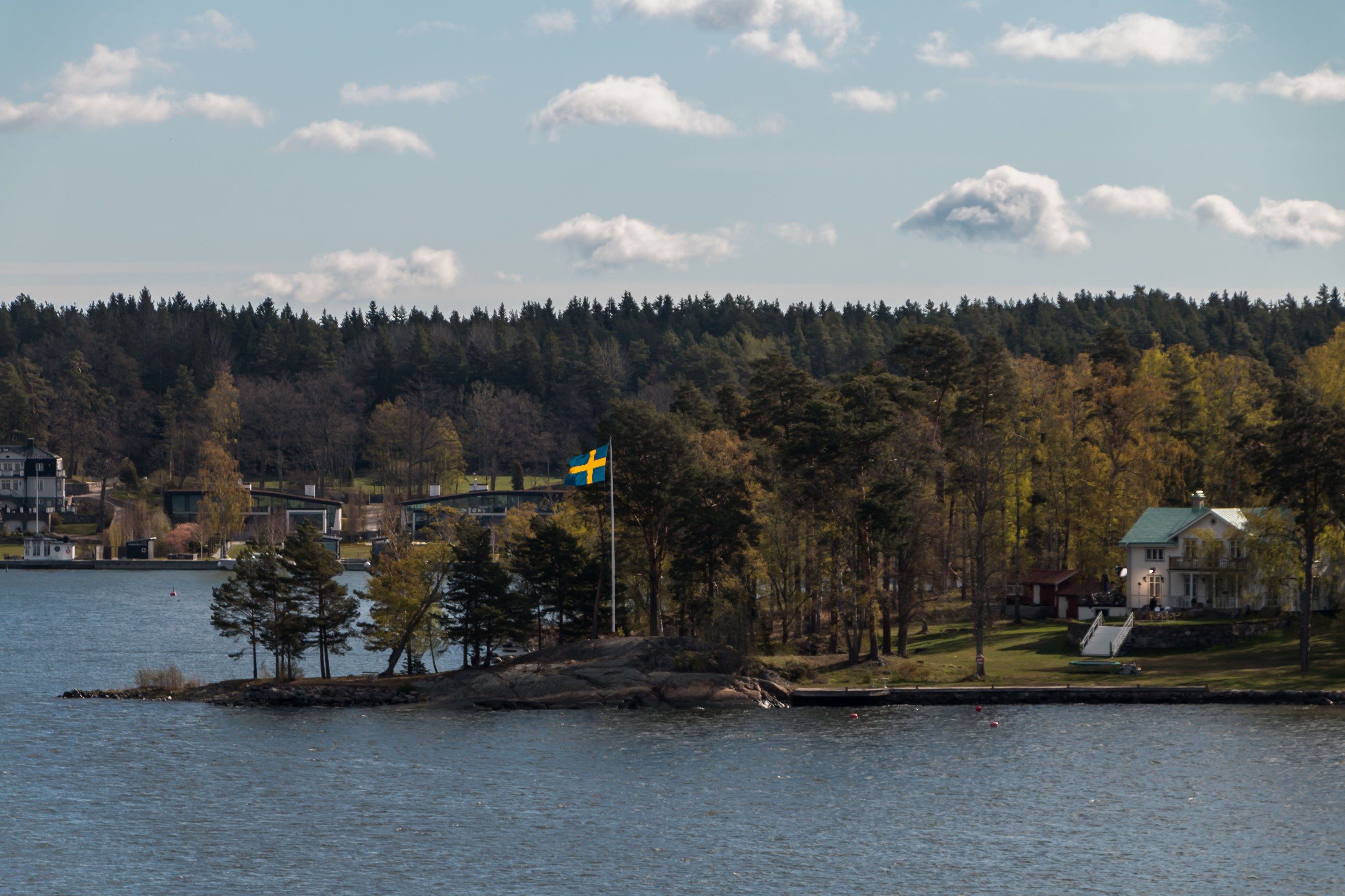 Hus vid svensk skärgård med blågul flagga och tallskog i bakgrunden.